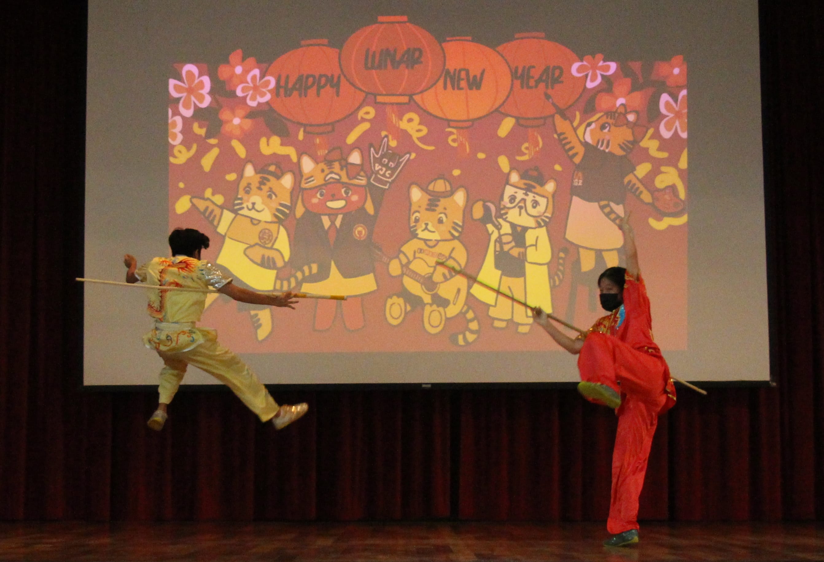 Two performers in colorful costumes do a martial arts staff routine on stage before a Happy Lunar New Year backdrop.